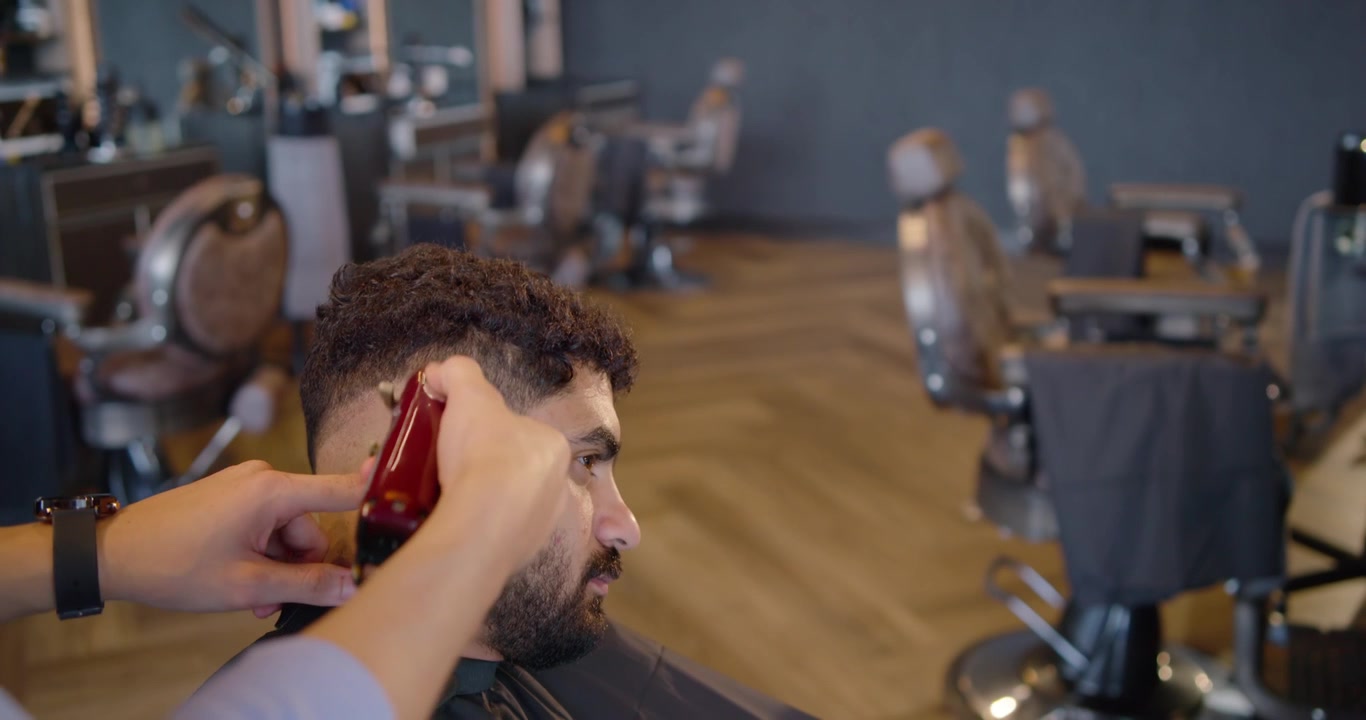 Barbeiro começando a cortar a nuca de cliente masculino na barbearia, usando aparador vermelho para modelar um degradê cônico, com espaço para texto.