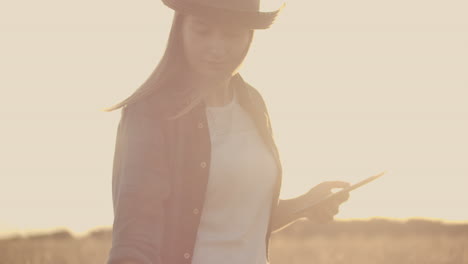 Una-Campesina-Con-Sombrero-Y-Camisa-A-Cuadros-Toca-Los-Brotes-Y-Las-Semillas-De-Centeno,-Examina-E-Ingresa-Datos-En-La-Tableta-Que-Se-Encuentra-En-El-Campo-Al-Atardecer.