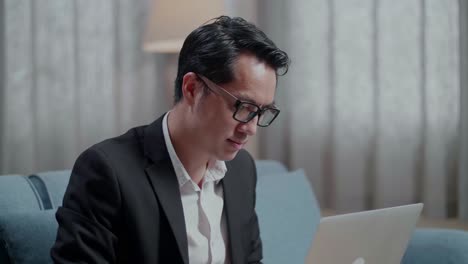 primer plano de un hombre de negocios asiático con chaqueta y pantalones cortos mirando a la cámara y sonriendo mientras trabaja con una computadora portátil en casa