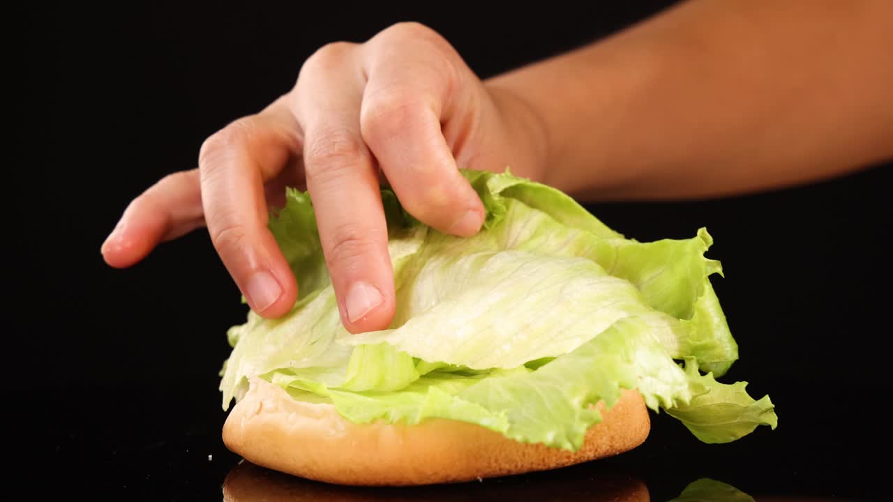 A hand carefully places and arranges fresh iceberg lettuce onto a burger bun against a black background, using even studio lighting and smooth camera movement