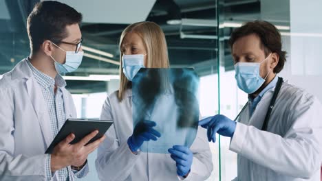 caucasian male and female doctors in medical masks speaking and discussing xray scan and typing on tablet device in hospital office