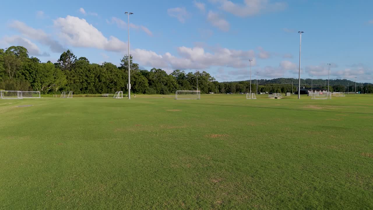Aerial footage captures an empty soccer field under clear skies, showcasing goalposts and lush greenery in the background