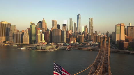 AERIAL:-Flight-over-Brooklyn-Bridge-with-American-flag-waving-and-East-River-view-over-Manhattan-New-York-City-Skyline-in-beautiful