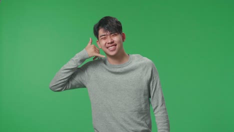 a cheerful asian man making a phone call hand gesture while standing in front of green screen background