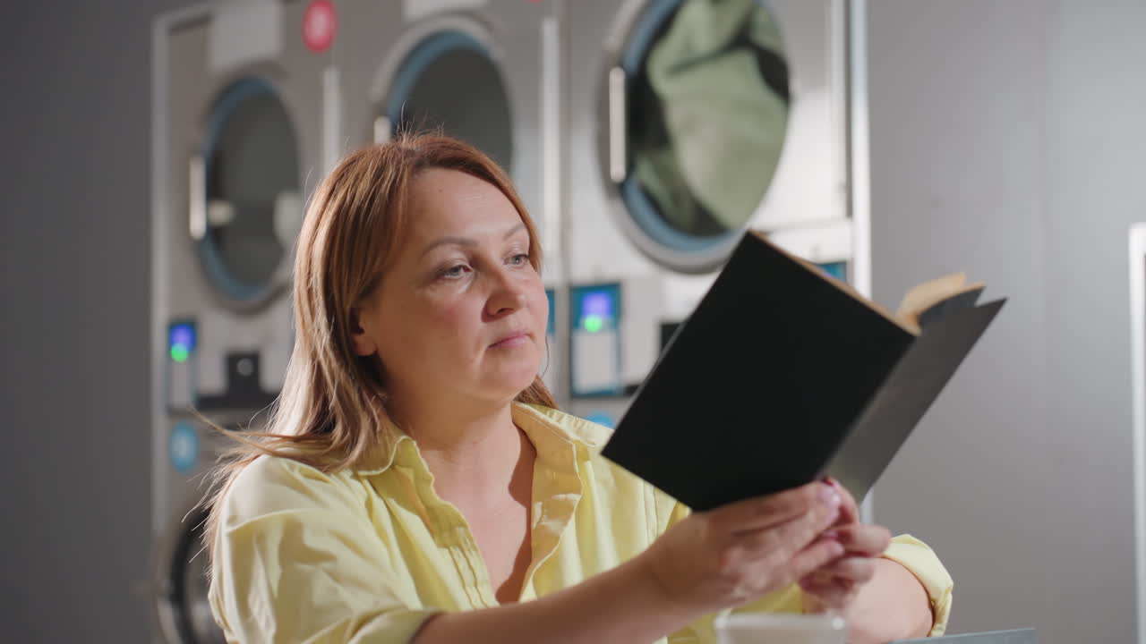 Female operator seated inside laundromat opens record book, smiles while reading, nods in approval with satisfaction, cup of coffee resting on steel table near industrial washing machines