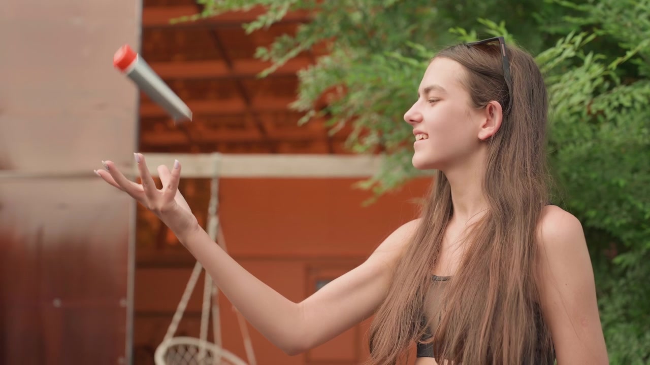 Una joven de cabello largo, con una blusa negra, interactúa al aire libre, gesticulando y mirando a su alrededor. El entorno incluye vegetación, una fachada de edificio naranja, una silla colgante, y una luz natural suave y tranquila.