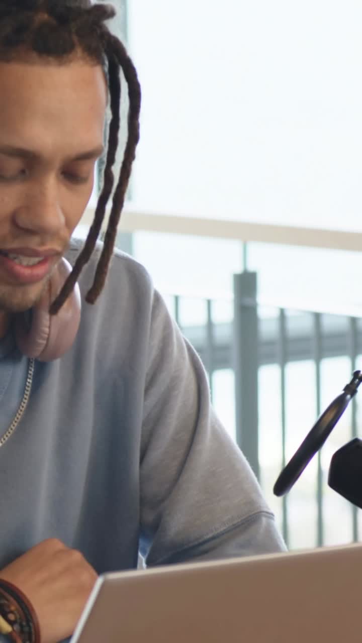 Vertical video: Looking at laptop, man typing, glancing at microphone, creating podcast at table