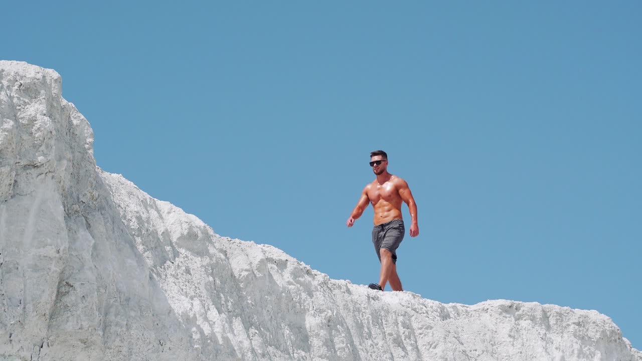 Bodybuilder athlete with a naked body in sunglasses and shorts is walking on a white mountain against a blue sky. Outdoor training