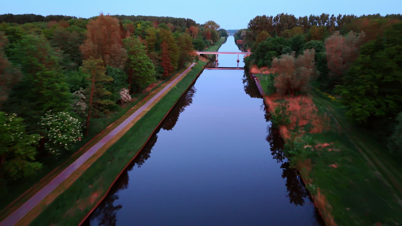 Flying over the mirror canal with a bridge above it. Deep woods grow around the canal. Sunset time footage.
