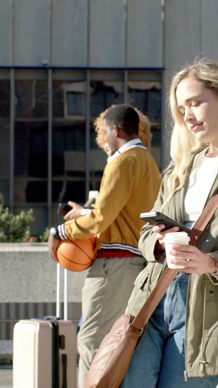 Vertikal video: Går på en solfylt plass, mann i jakke med basketball og veske, kvinne sjekker telefonen.