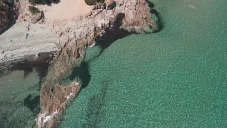 Vista-Panorámica-De-Las-Olas-Del-Mar-Tropical-Y-Cristalino-Que-Se-Estrellan-Contra-Las-Rocas-De-La-Costa-En-Cerdeña,-Italia---Toma-Estática-Y-Aérea-De-Drones