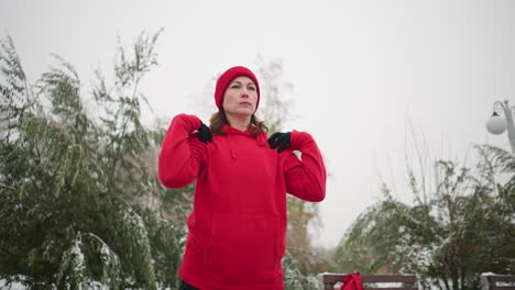 lady performing hand stretch by turning her arm in a fitness routine outdoors, dressed in athletic wear, background featuring frosted tree, bag on bench and snow everywhere