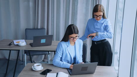 female colleagues in office looking at laptop discussing work. blonde woman standing and looking at her watch and then at computer again.