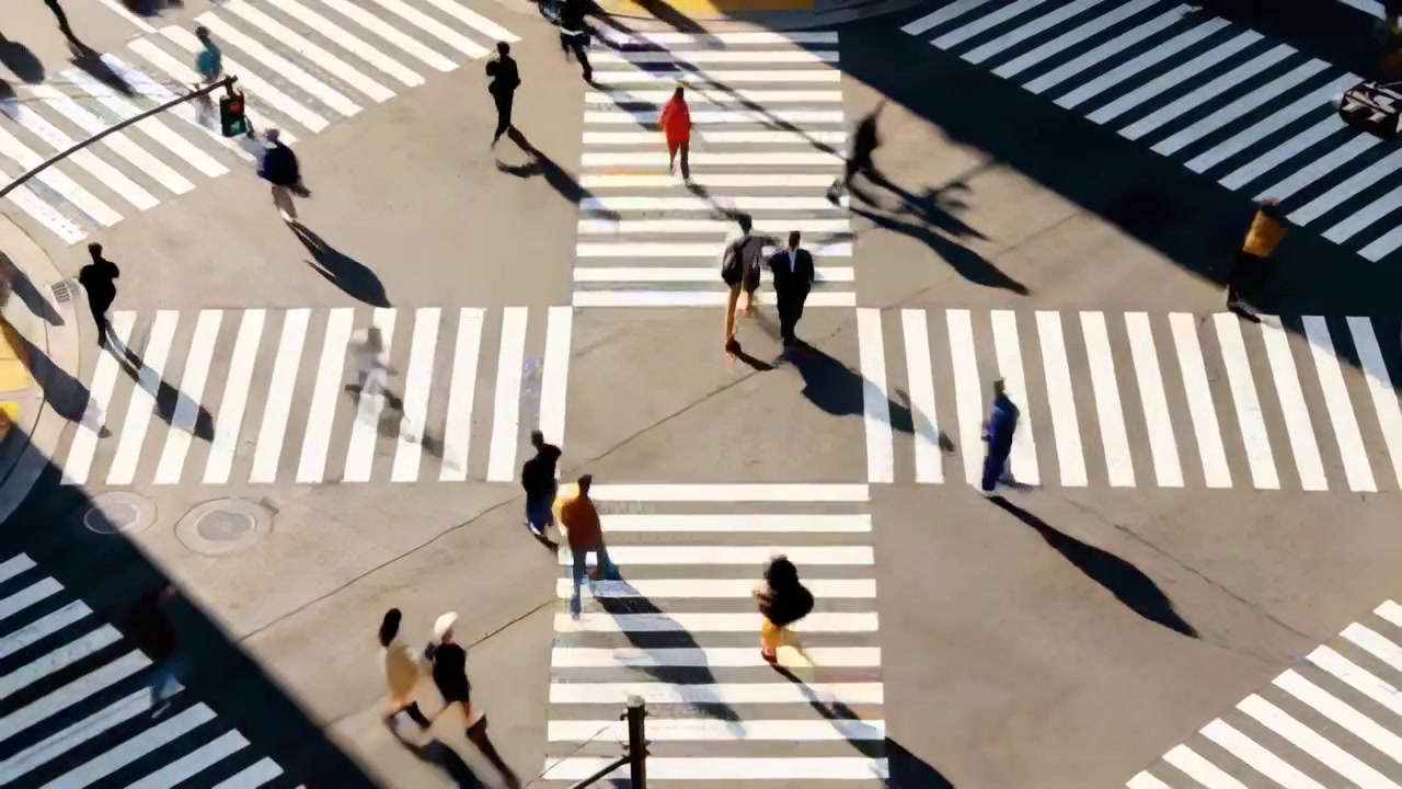 Personas cruzando una intersección, con el semáforo en verde, abrigos de colores vivos, pasos de cebra y sombras.