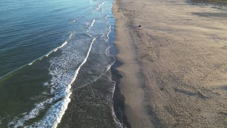 Vuelo-De-Drones-En-Una-Playa-Con-Suaves-Olas-Dividiendo-El-Rodaje-En-El-Lado-Izquierdo-El-Mar-Y-En-El-Lado-Derecho-La-Playa-Con-La-Arena-Y-La-Gente-Caminando-En-Una-Tarde-De-Invierno-En-Valencia-España