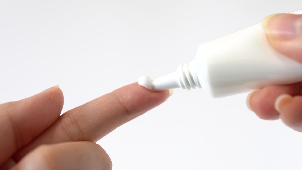 vista lateral de primer plano de las manos de la dama aplicando crema hidratante cayendo sobre un dedo en fondo blanco