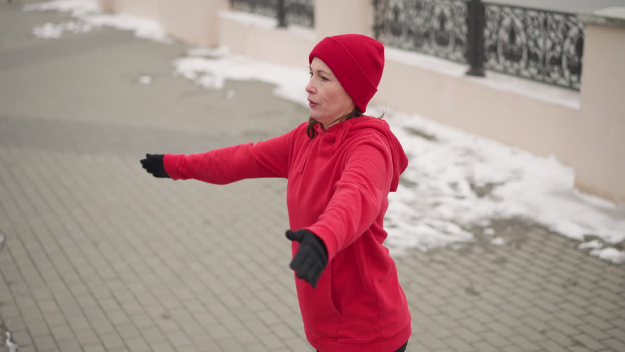 Woman in red hoodie and gloves performing hand stretches outdoors on snow-covered path with decorative metal fence in winter environment in cold weather atmosphere
