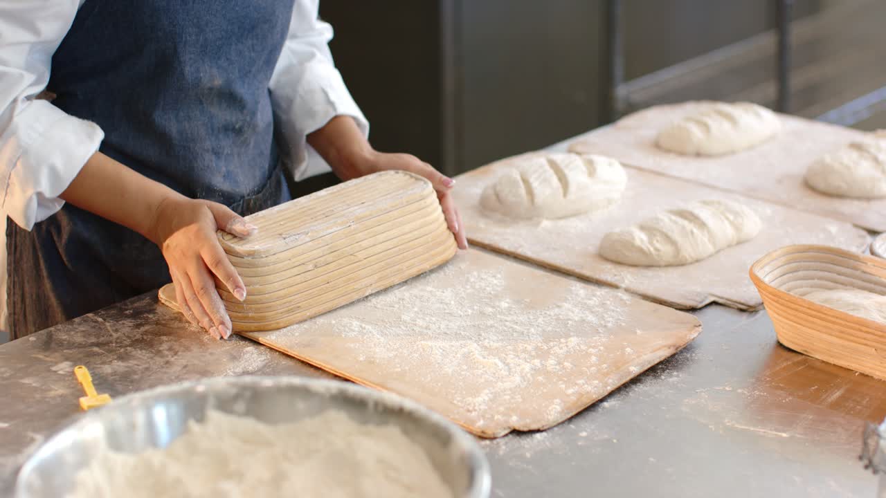 Mulher afro-americana de avental em uma loja, levantando um banneton e soltando um pão em uma tábua para moldar antes de ir ao forno