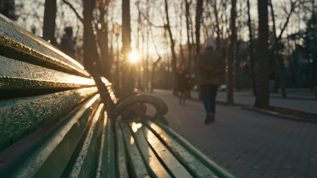 Golden light reflects on Central Park green bench as the sun sets. People walking on background. Cold weather in late autumn. Chisinau Moldova