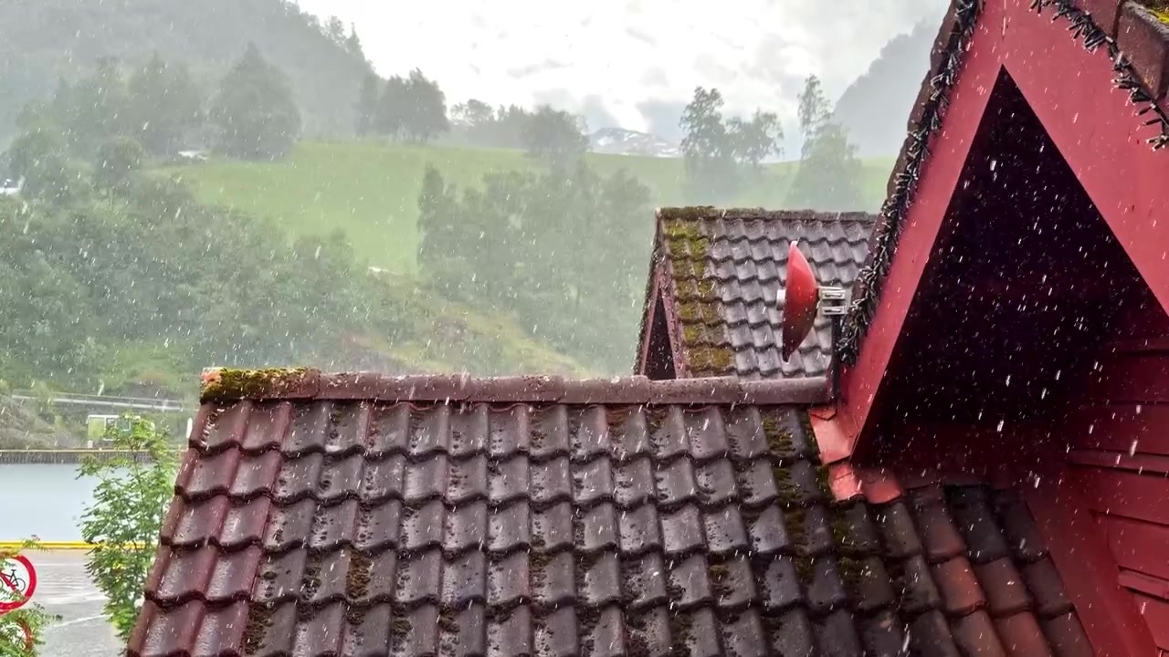 Heavy rain falls on traditional red-tiled roof with green hills in background