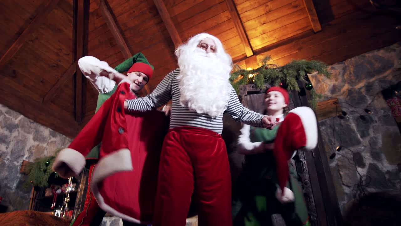 Santa doing exercises at home. Elves help Santa to put on his costume. Old bearded Santa Claus dresses his red suit at Christmas eve. View from below