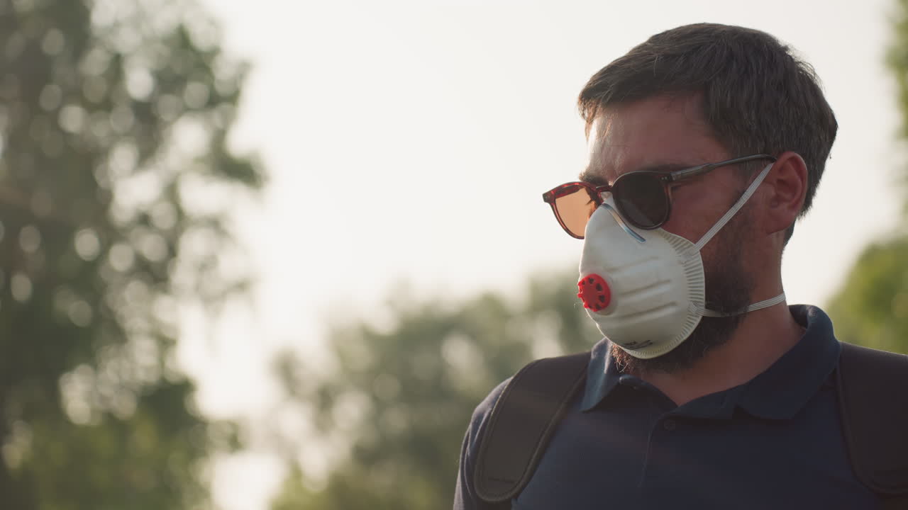 Close up of man wearing nose mask and sunglasses gazing intently at field with sunlight filtering through trees, beard visible, suggesting careful inspection of crops and safety preparedness