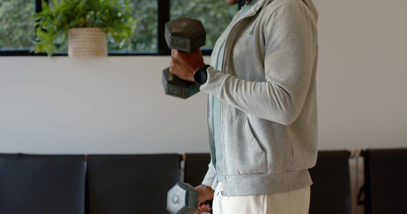 Uomo afroamericano con felpa con cappuccio che fa curl per i bicipiti con manubri esagonali nella palestra di casa per il fitness.