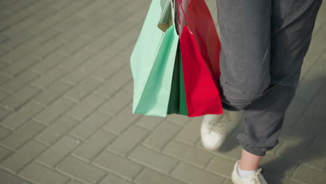 leg view of individual wearing grey joggers and white canvas sneakers, holding green, mint, and red shopping bags, walking casually on an interlocked path in an urban setting