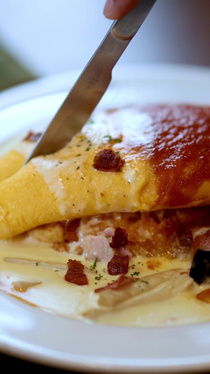 Primo piano di un coltello che taglia l'omurice, con salse cremose e al pomodoro, luce naturale, profondità di campo ridotta.