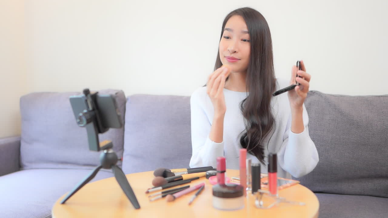 비디오 모드에서 스마트폰 앞에 있는 한 젊은 미녀가 소셜 미디어 메이크업 자습서 중간에 있습니다.