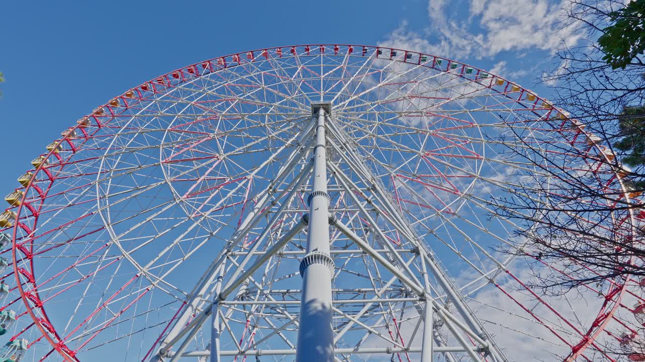 Wide low-angle view showing the full scope of a massive white and red Ferris wheel reaching into a bright blue sky