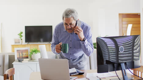 Video-of-senior-biracial-man-talking-on-smartphone,-drinking-coffee-and-using-laptop-at-home