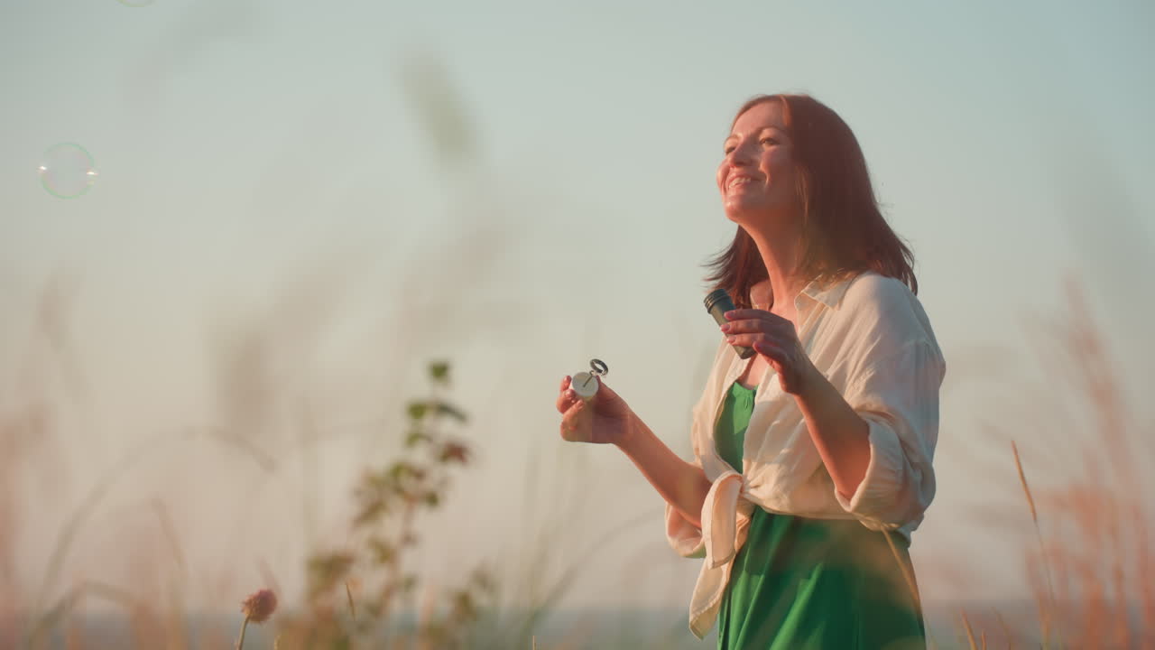 Fair skinned woman in green dress standing cliff edge at river valley sunset, smiling blowing iridescent soap bubbles floating gently over grasses and water