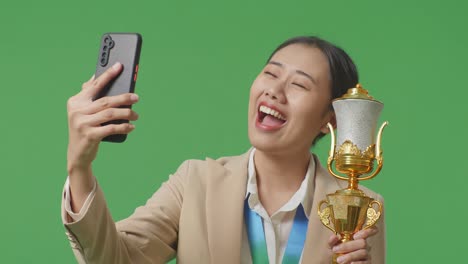 close up of asian business woman in a suit with a gold medal showing a gold trophy to the video call on smartphone on green screen background in the studio