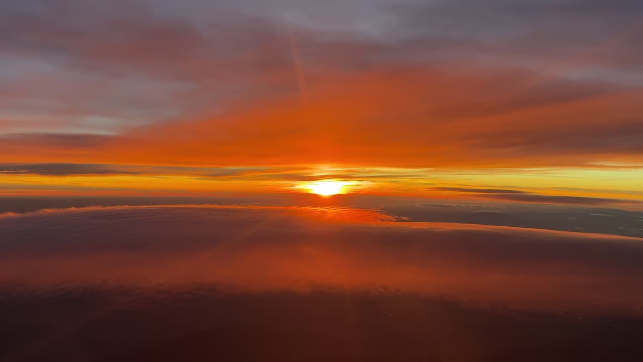 A pilot’s eye view from the cockpit of a jet flying in the upper atmosphere at sunset between layers of ethereal orange color clouds with the sun veiled in the horizon