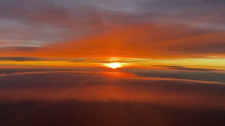 La prospettiva di un pilota dalla cabina di pilotaggio di un jet che vola nell'atmosfera superiore al tramonto tra strati di nuvole eteree color arancio, con il sole velato all'orizzonte