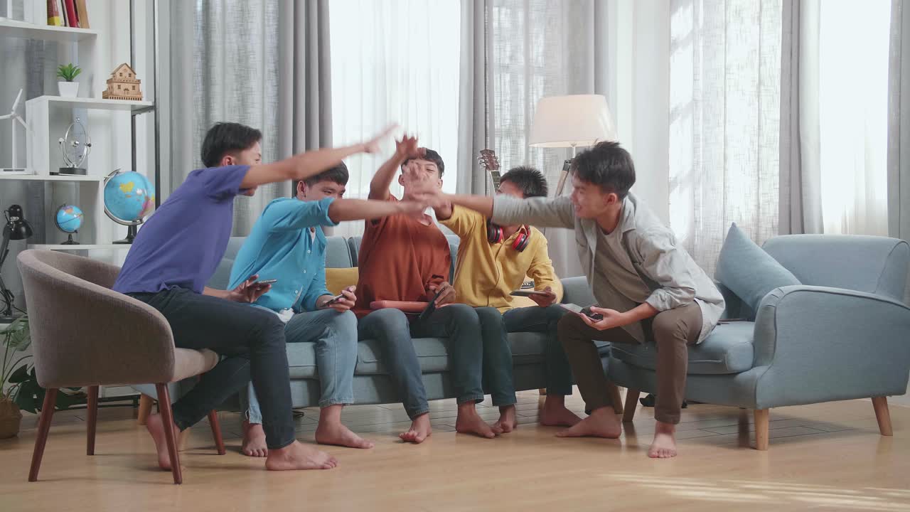 음악 악기를 가진 젊은 아시아 친구 그룹이 집에서 소파에서 스마트폰에서 게임을 하기 전에 손을 잡고 있습니다.