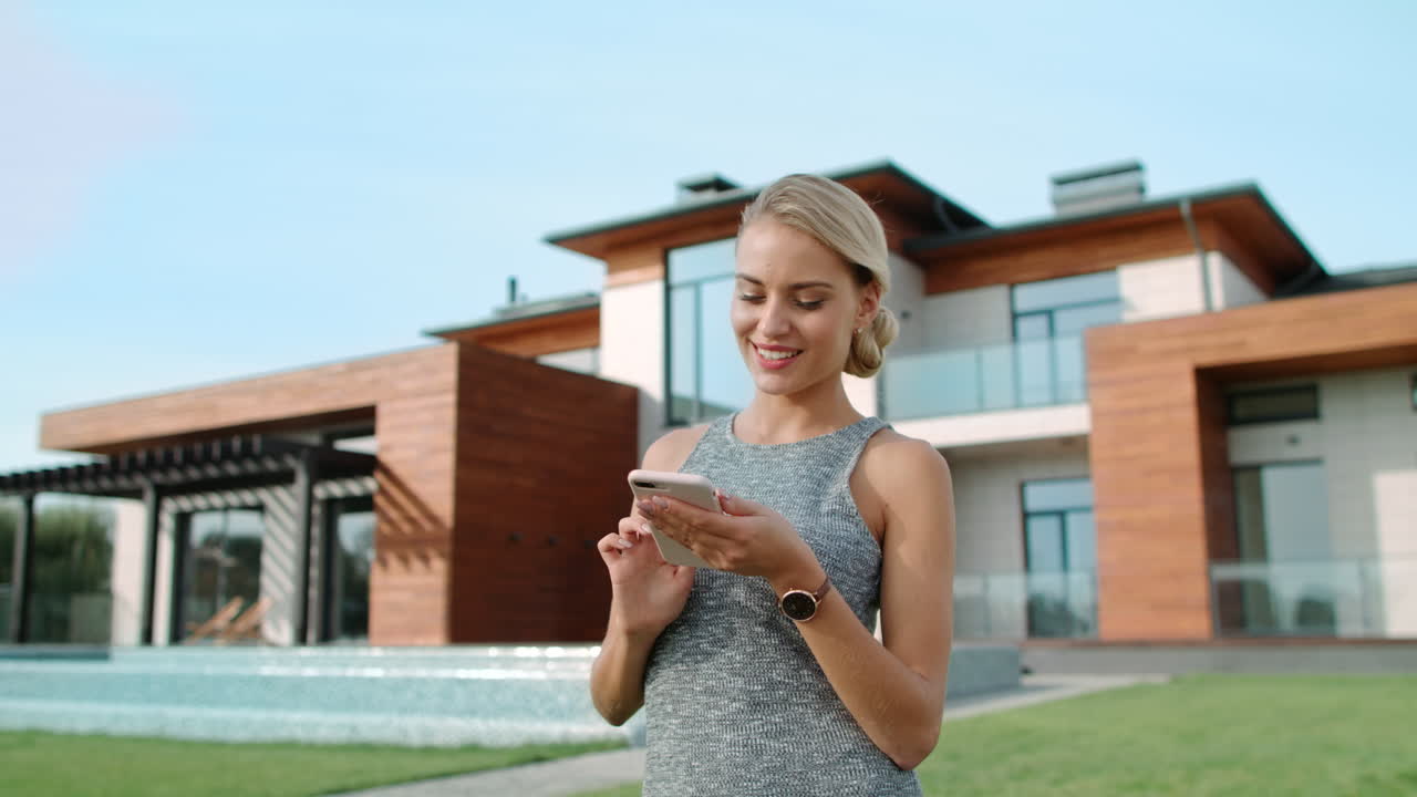 Beautiful woman typing mobile near luxury house exterior. Successful lifestyle