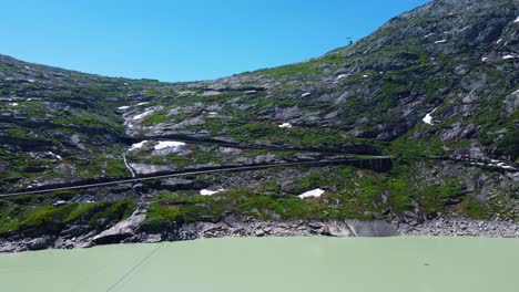 Aerial-drone-shot-over-a-winding-road-beside-a-waterfall-in-distance-in-Grimselpass-high-mountain-road-along-beautiful-Swiss-Alps,-Switzerland-at-daytime