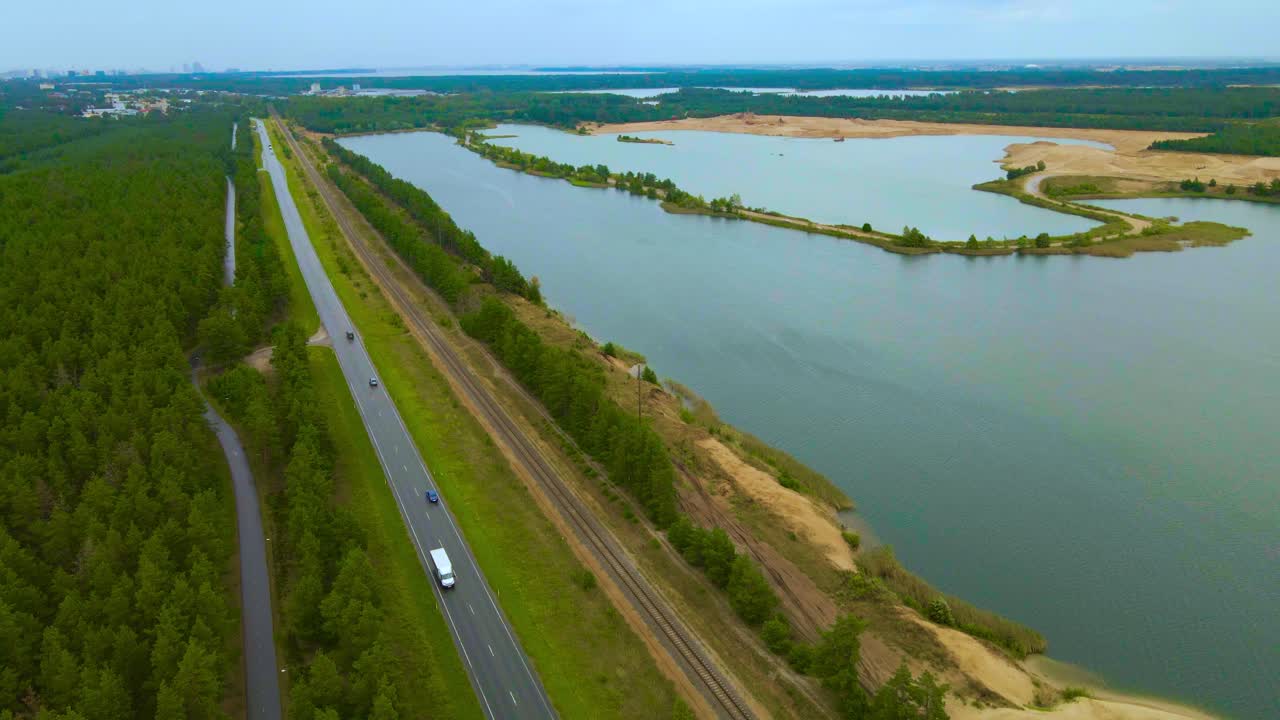 Aerial drone footage flying above a large open pit sand quarry mine during a cloudy day while a highway with cars is visible next to a large dug and excavated artificial lake. Mining roads visible