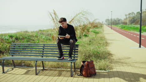 man picking up notes from the backpack and starting to study on the park bench
