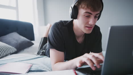 Estudiante-Universitario-Masculino-Con-Audífonos-Trabaja-En-La-Cama-En-Una-Casa-Compartida-Con-Una-Computadora-Portátil