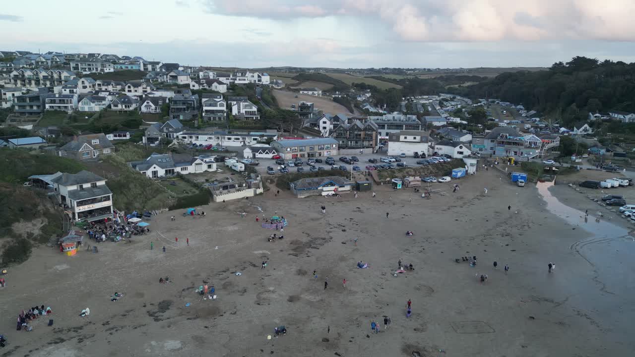 Drone video captures Polzeath Beach, Cornwall, at dusk, showcasing a lively seaside town with people enjoying the sandy shores, surrounded by charming coastal architecture