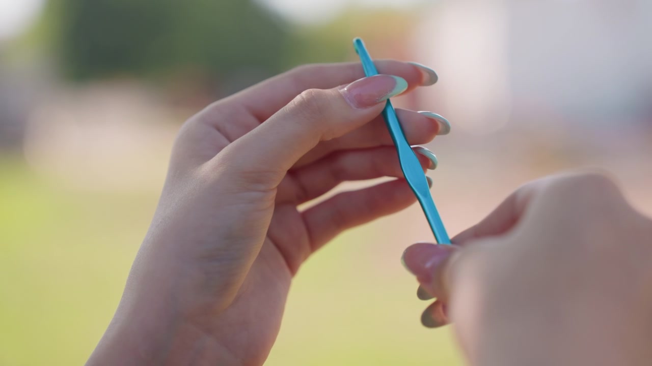 Primo piano di mani con unghie lunghe che tengono un uncinetto blu, dimostrando la tecnica dell'uncinetto. Le mani manipolano l'attrezzo e il filato in una luce naturale soffusa, concentrandosi sui movimenti dettagliati delle mani.