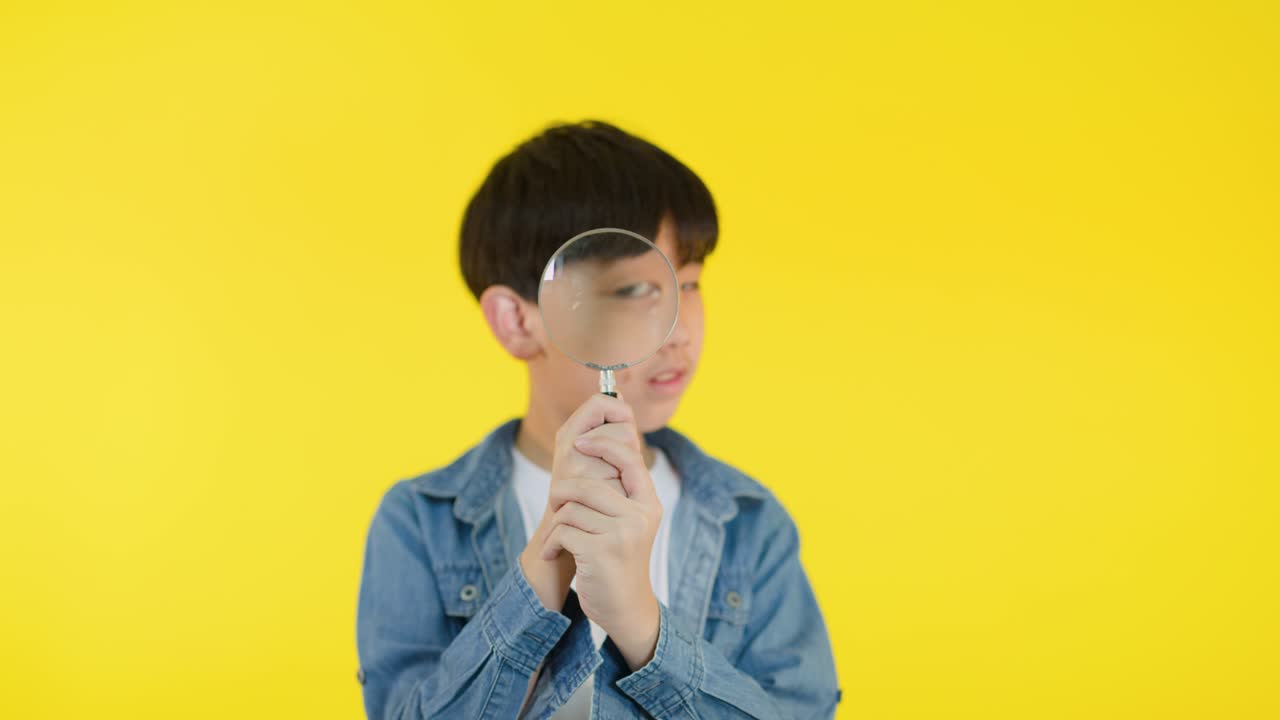 Smiling child in denim jacket explores with magnifying glass, bright yellow backdrop, playful close-up