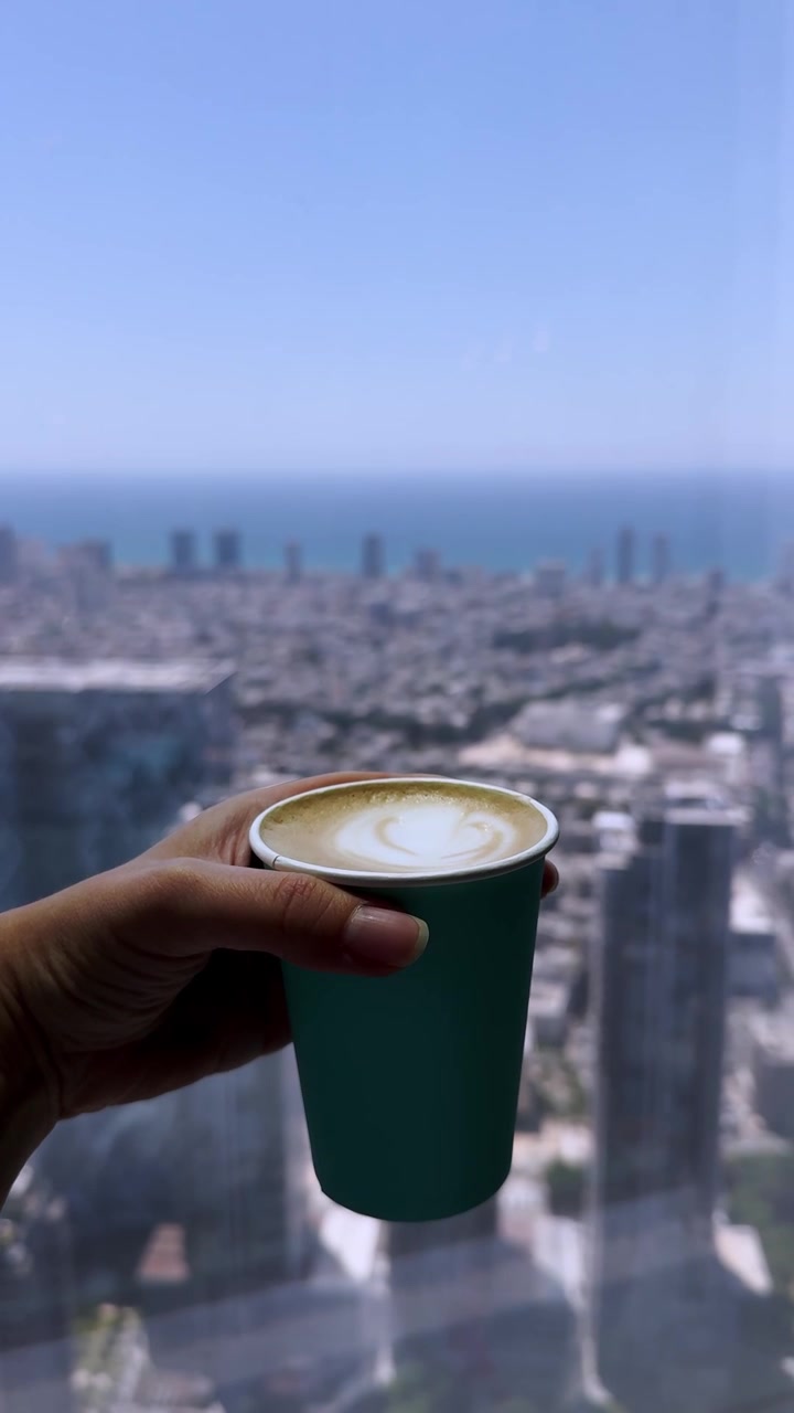 Mão segurando uma xícara de cappuccino em um edifício alto com vista para a cidade.