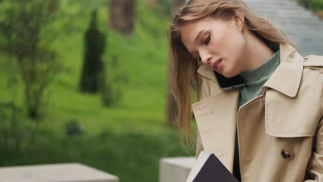 estudante caucasiana a falar ao telefone enquanto escreve num caderno ao ar livre.