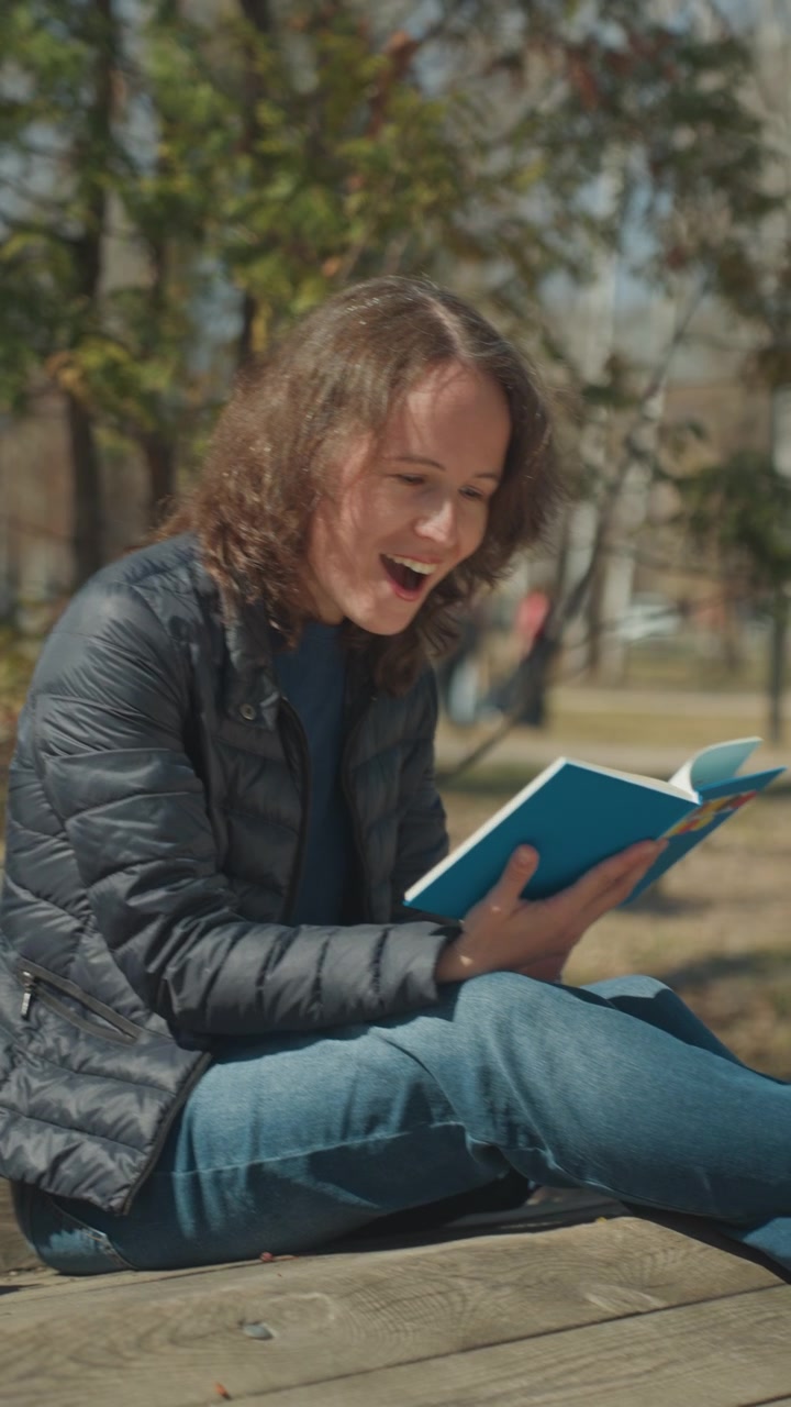 Close up of woman reading blue book outdoors on wooden bench with trees and park in background. Book is open held by hands pages visible. Soft natural light creates clear shadows on subject