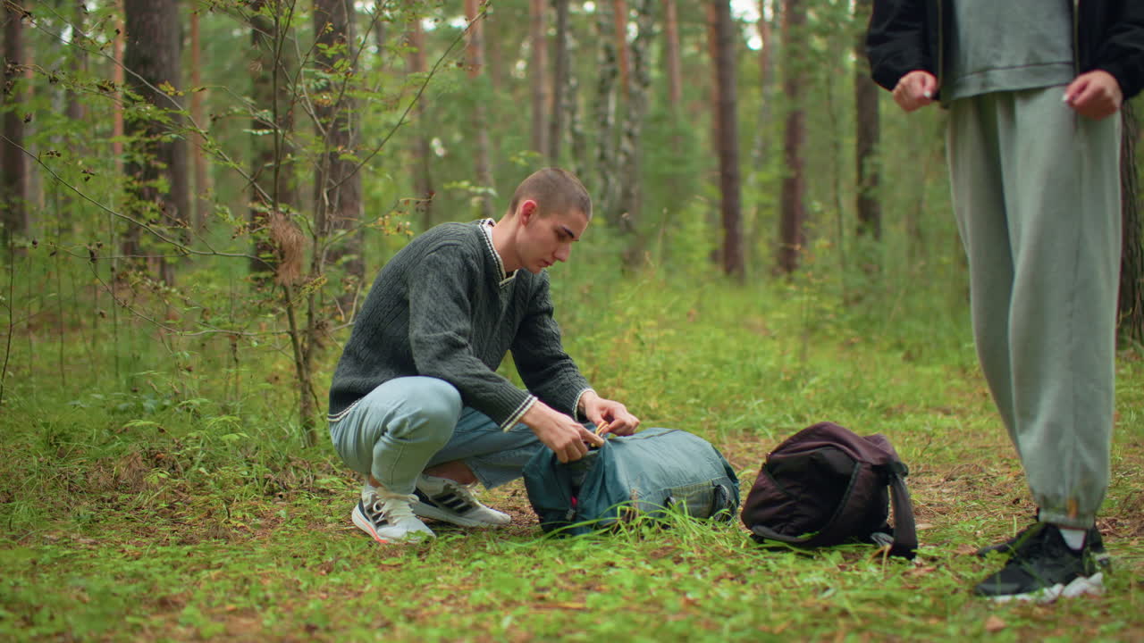 young man crouching in green forest carefully unzipping backpack while another person stands partially visible nearby, surrounded by trees and grass in calm woodland setting