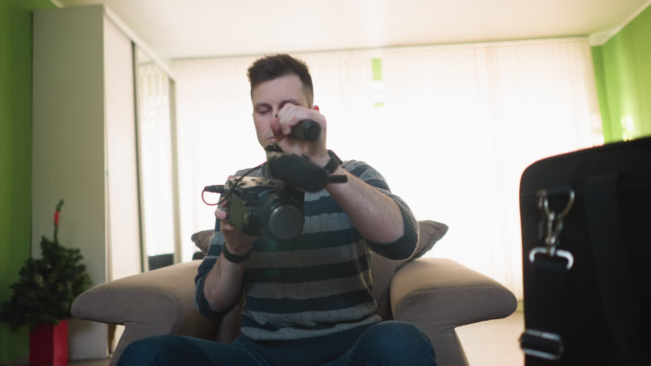 Homem ajustando câmera portátil com microfone em ambiente de estúdio, focado nas configurações da câmera. Ambiente interno confortável com paredes verdes e sofá ao fundo. Preparando-se para produção de vídeo.